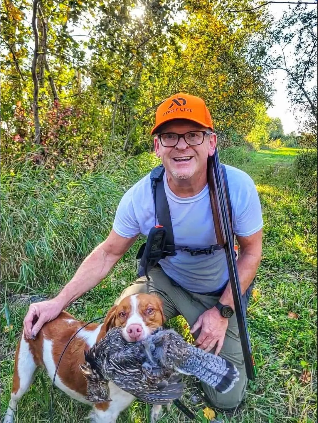 Cory Fechner kneeling on a grassy trail in blaze orange, his Brittany delivering a ruffed grouse to hand